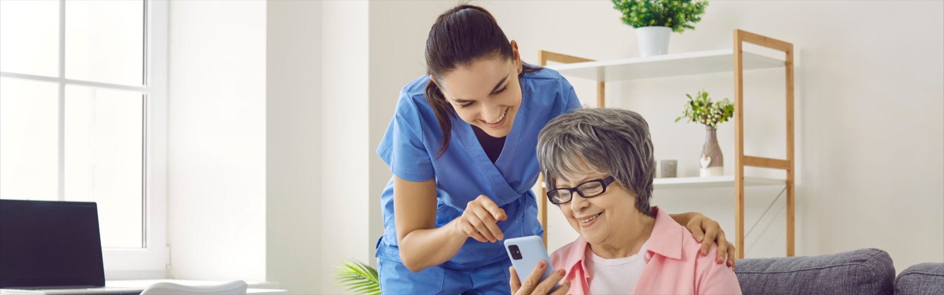 Smiling caregiver and senior woman in nursing home looking at mobile phone screen together.