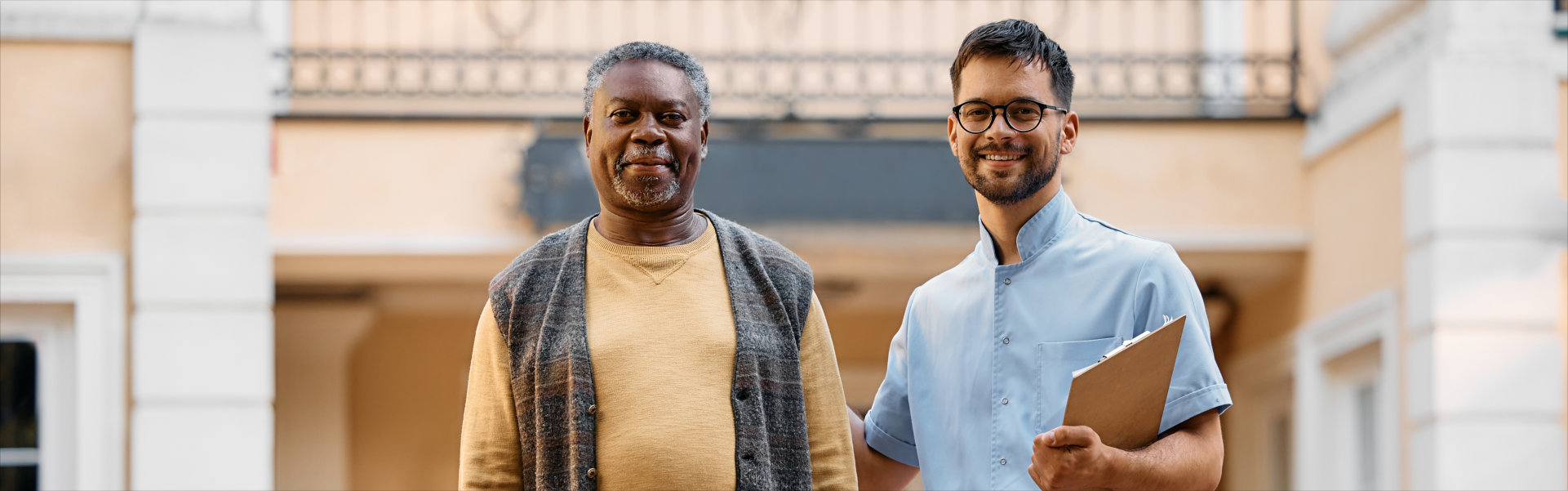 Smiling black senior man and young caregiver in front of residential care home looking at camera.